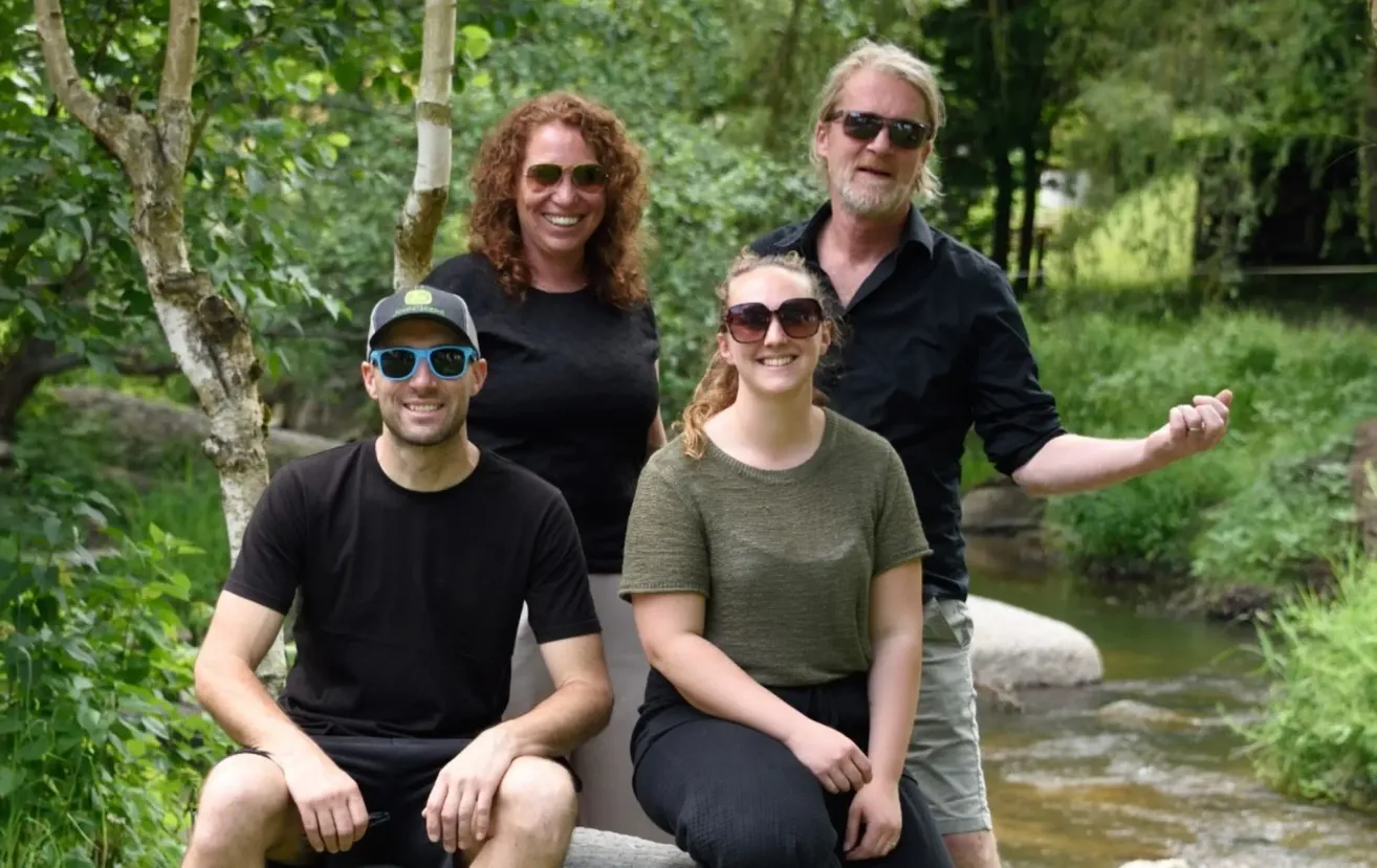 Vier Menschen lächeln für ein Foto in der Nähe eines Flusses. Sie stehen oder sitzen auf Felsen und tragen Sonnenbrillen. Bäume und Grünflächen sind im Hintergrund.