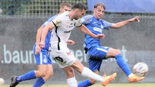 Drei Fußballspieler sind auf einem nassen Feld in Aktion. Zwei sind in blauen Trikots, einer in weiß, und sie konzentrieren sich alle auf den Ball.