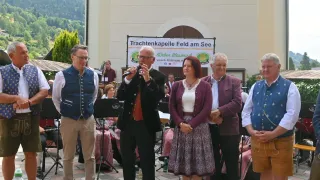 Vier Personen stehen vor einer Kirche, mit einem Banner, das Trachtenkapelli Feld am See besagt. Ein Mann spricht in ein Mikrofon. Dahinter spielt eine Band.