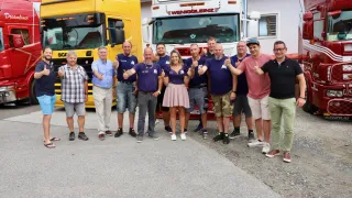 Eine Gruppe von Menschen posiert für ein Foto vor zwei Lastwagen. Der Mann in der Mitte trägt eine pinke Rock. Die Lastwagen tragen die Namen Gomschak und Weniggleinz.