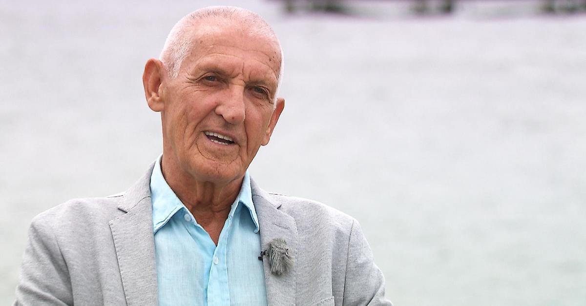 Ein älterer Mann mit weißen Haaren, der ein hellblaues Hemd und einen grauen Blazer trägt, lächelt im Freien.