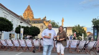 Ein Mann und eine Frau stehen vor einer Outdoor-Kino-Aufstellung mit einem Publikum in Stühlen, vor der Kulisse eines historischen Gebäudes.