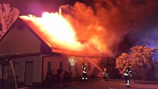 Feuerwehrleute bekämpfen nachts einen Hausbrand, wobei die Flammen das Dach verschlingen und starker Rauch aufsteigt. Drei Feuerwehrleute spritzen Wasser aus einem Schlauch.