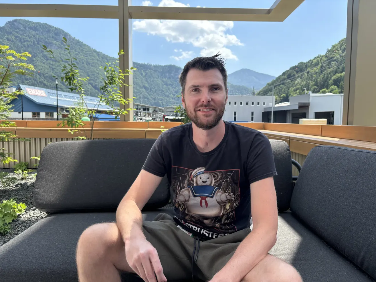 Ein Mann sitzt auf einer Couch mit einem lächelnden Gesicht. Er trägt ein schwarzes T-Shirt mit einem gedruckten Design. Dahinter befinden sich Berge, ein Gebäude und ein klarer blauer Himmel.