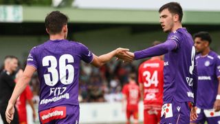 Zwei Fußballspieler in lila Trikots schütteln sich auf dem Spielfeld die Hände. Der Spieler auf der linken Seite hat die Nummer 36, während der Spieler auf der rechten Seite die Nummer 34 hat. Sie schauen beide nach vorne.