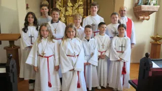 Eine Gruppe von Kindern in weißen Gewändern mit roten Schärpen und Kreuzen steht in einer Kirche. Dahinter stehen zwei Priester.