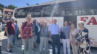 Eine Gruppe von Menschen steht an einem sonnigen Tag vor einem Bus. Der Bus zeigt die Reflexion eines Gebäudes an seiner Seite. Die Gruppe besteht aus älteren Personen, eine trägt einen Hut, und sie scheinen auf etwas zu warten.