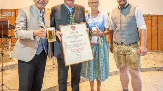 Vier Personen in traditioneller Kleidung, möglicherweise bei einer Preisverleihung. Der Mann in der Mitte hält eine gerahmte Urkunde. Der Mann auf der linken Seite hält ein Glas Bier und eine Trophäe. Die Frau auf der rechten Seite hält einen weißen Becher. Der Mann auf der rechten Seite trägt eine graue Weste und braune Hose.
