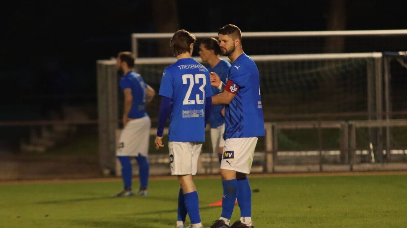Eine Gruppe von Fußballspielern in blauen Trikots und weißen Shorts steht auf einem Rasenfeld. Zwei Spieler stehen sich gegenüber und unterhalten sich vielleicht. Im unscharfen Hintergrund befindet sich ein Zaun und ein Fußballtor.