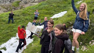 Eine Gruppe von Wanderern, einschließlich eines Mannes und Frauen, ist auf einem Berghang unterwegs, trägt eine Kiste und posiert für ein Foto. Einige tragen Brillen, Mützen und Rucksäcke.