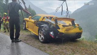 Ein gelber Sportwagen liegt auf der Straße, mit sichtbaren Schäden an der Front und hinten. Er wird von einem Abschleppwagen gezogen. Das Auto ist mit Pflanzen bedeckt. Im Hintergrund befinden sich Berge und ein bewölkter Himmel.