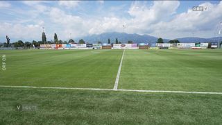 Ein leerer Fußballplatz mit grünem Rasen und weißen Markierungen. Werbebanner sind am Zaun angebracht. Berge und Bäume sind im Hintergrund.