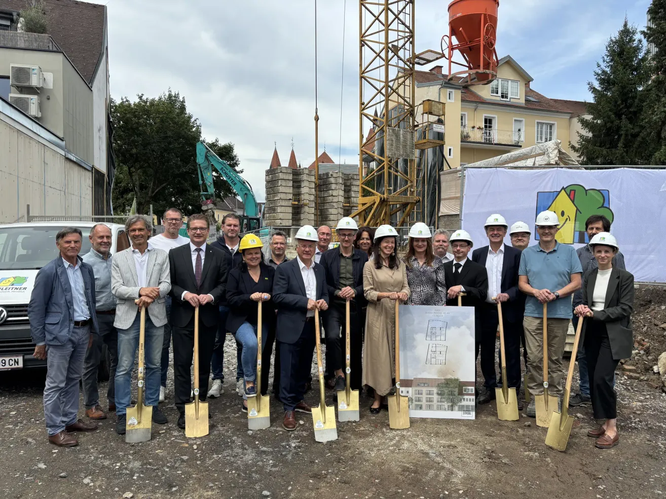 Eine Gruppe von Menschen in Schutzhelmen posiert für ein Foto an einer Baustelle. Sie halten Schaufeln und stehen vor einem Banner und einem Gebäude im Bau. Dahinter steht ein Kran und mehrere Häuser.