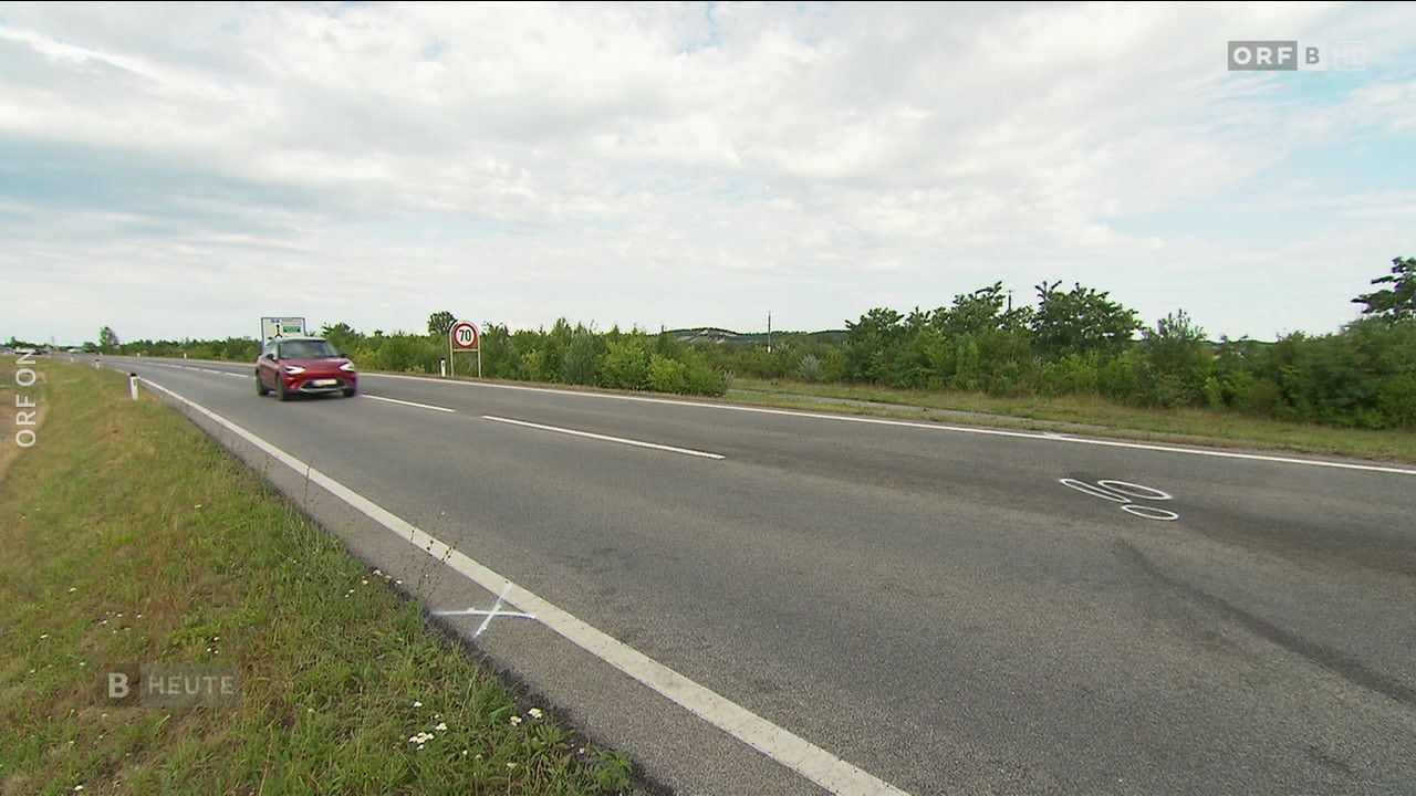 Ein rotes Auto fährt auf einer zweispurigen Straße. Das Geschwindigkeitsschild zeigt 70 km/h an. Die Straße ist von grünem Gras und Bäumen umgeben.