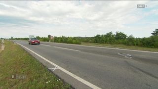 Ein rotes Auto fährt auf einer zweispurigen Straße. Das Geschwindigkeitsschild zeigt 70 km/h an. Die Straße ist von grünem Gras und Bäumen umgeben.