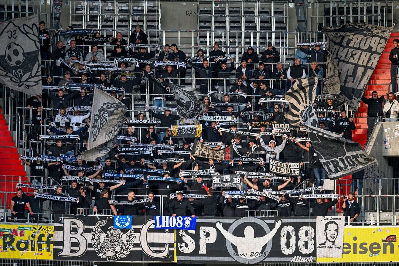 Fans in einem Stadion halten Banner und Fahnen hoch. Viele von ihnen tragen schwarze Kleidung, und die Menge ist groß. Ein großes Banner mit einem Logo und der Nummer 08 ist sichtbar.