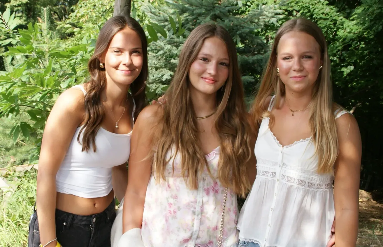 Drei junge Frauen stehen eng beieinander in einem Garten und lächeln für ein Foto. Eine trägt ein weißes ärmelloses Top, eine andere eine Blumenbluse und die dritte ein weißes Oberteil mit Jeansshorts. Sie haben lange Haare und sind mit Halsketten geschmückt.