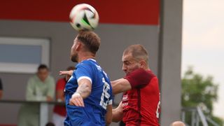 Zwei Fußballspieler konkurrieren, einer in Blau, der andere in Rot. Der Spieler in Blau balanciert einen Ball auf seinem Kopf. Eine Person in Rot streckt die Hand aus, um den Ball zu berühren.