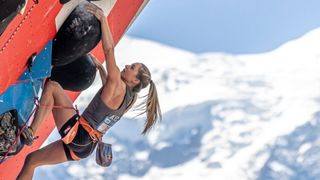 Eine kletternde Frau mit Pferdeschwanz hält sich mit den Händen an einem Klettergriff fest und trägt ein graues Tanktop mit der Nummer 17.
