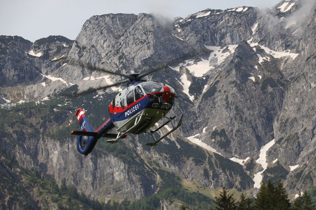 Ein Polizeihubschrauber mit roten, weißen und blauen Markierungen fliegt über eine Bergregion mit verschneiten Gipfeln und grünen Tälern.