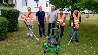 Fünf Personen stehen auf einem Rasen und lächeln für ein Foto. Sie tragen orange Westen und halten verschiedene Werkzeuge. Der Rasenmäher in der Mitte ist grün. Hinter ihnen ist ein Baum und ein Gebäude mit Fenstern zu sehen.