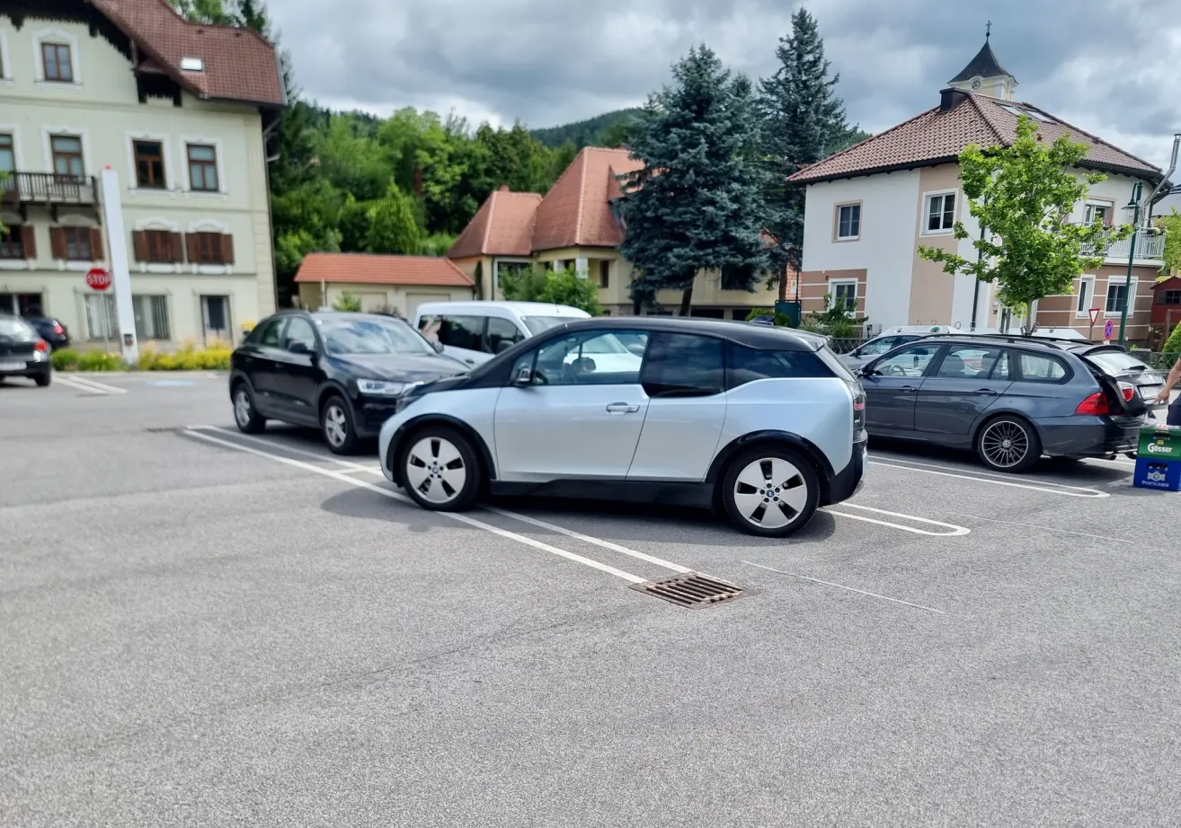 Ein hellblaues Auto steht in einem Parkplatz neben zwei anderen Autos. Die Gebäude hinter den Autos haben braune Dächer.