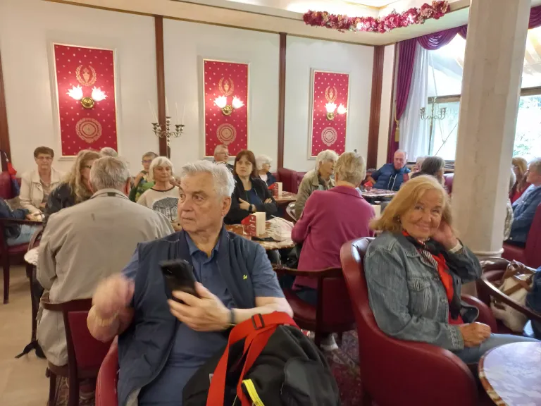 Eine Gruppe älterer Erwachsener versammelt sich in einem Restaurant. Ein älterer Mann benutzt sein Handy. Eine Frau in der Nähe lächelt. Es gibt rote Stühle und Tische, und die Wände sind mit roten dekorativen Elementen geschmückt.