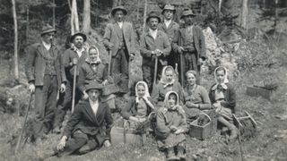 Ein Schwarz-Weiß-Gruppenfoto einer Gemeinschaft, möglicherweise aus einem ländlichen Gebiet, steht und sitzt auf Gras mit Körben und Stöcken.