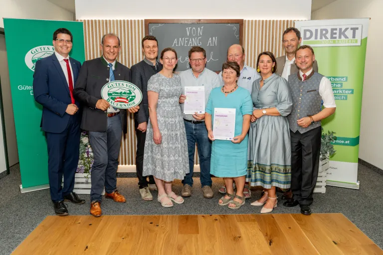 Eine Gruppe von Menschen in formeller Kleidung steht gemeinsam für ein Foto, einige halten Urkunden und ein Mann hält ein grünes Schild. Der Hintergrund zeigt eine Tafel und eine Wand mit Holzstreifen.