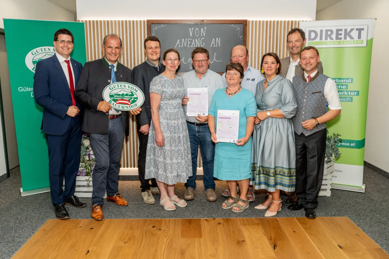 Eine Gruppe von Menschen in formeller Kleidung steht gemeinsam für ein Foto, einige halten Urkunden und ein Mann hält ein grünes Schild. Der Hintergrund zeigt eine Tafel und eine Wand mit Holzstreifen.