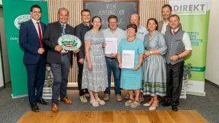 Eine Gruppe von Menschen in formeller Kleidung steht gemeinsam für ein Foto, einige halten Urkunden und ein Mann hält ein grünes Schild. Der Hintergrund zeigt eine Tafel und eine Wand mit Holzstreifen.