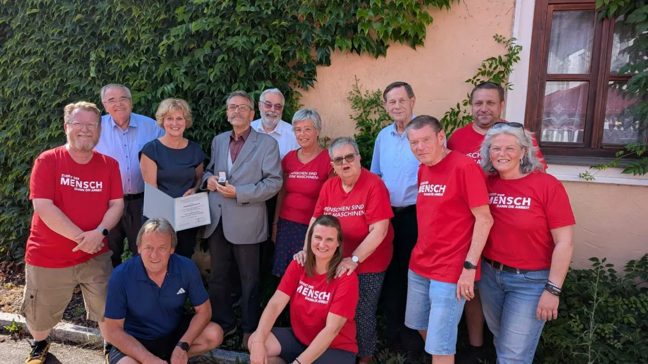 Eine Gruppe von Menschen posiert für ein Foto draußen; einige tragen rote T-Shirts mit Text und andere halten ein Zertifikat. Sie stehen vor einer Wand mit Pflanzen.