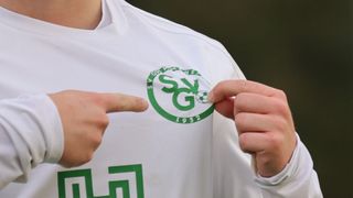 Eine Person in einem weißen Trikot mit einem grünen Logo und der Aufschrift 'SGV 1932' zeigt auf das Logo.