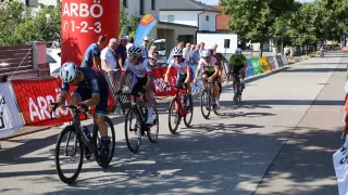 Eine Gruppe von Radfahrern konkurriert in einem Rennen, während mehrere Leute von der Seite zuschauen. Die Radfahrer tragen Helme und Radfahreranzüge. Im Hintergrund befindet sich ein roter Banner mit dem Wort 'Burgenland' darauf.