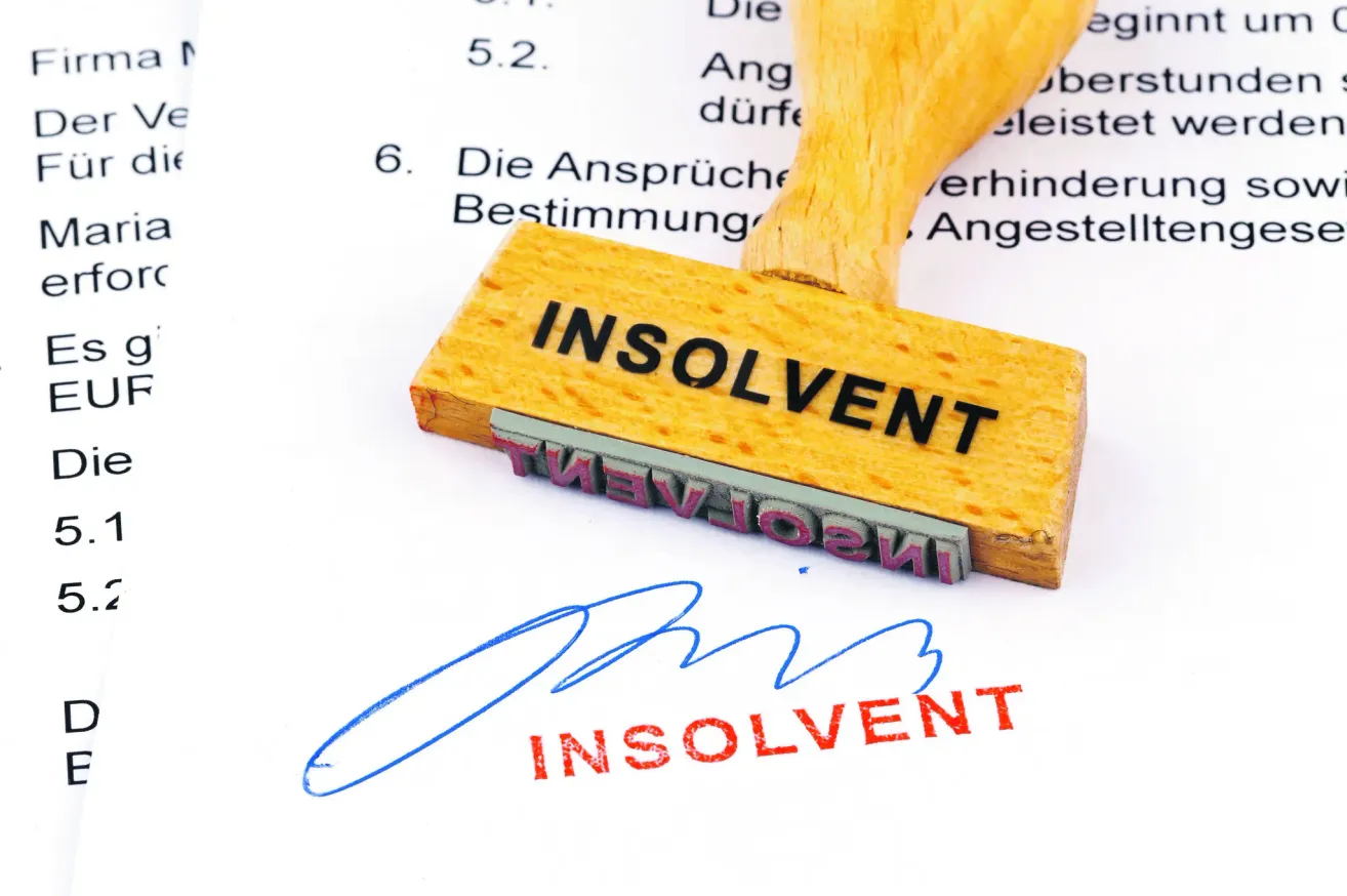Ein hölzerner Stempel mit schwarzer Schrift 'INSOLVENT' auf einem weißen Dokument mit einer blauen Unterschrift und verschwommenem deutschem Text.