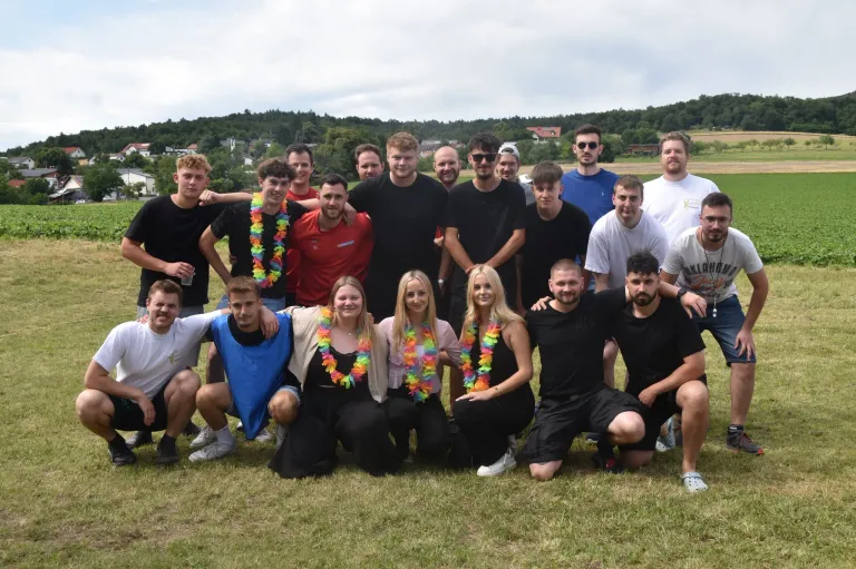 Eine Gruppe von Menschen in Freizeitkleidung posiert für ein Foto auf einem Grasfeld mit Bäumen und Gebäuden im Hintergrund.