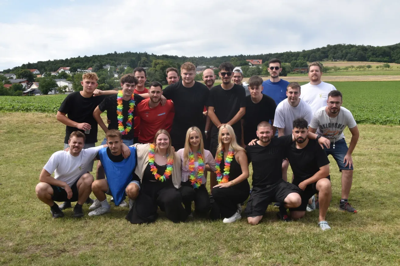 Eine Gruppe von Menschen in Freizeitkleidung posiert für ein Foto auf einem Grasfeld mit Bäumen und Gebäuden im Hintergrund.