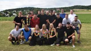 Eine Gruppe von Menschen in Freizeitkleidung posiert für ein Foto auf einem Grasfeld mit Bäumen und Gebäuden im Hintergrund.