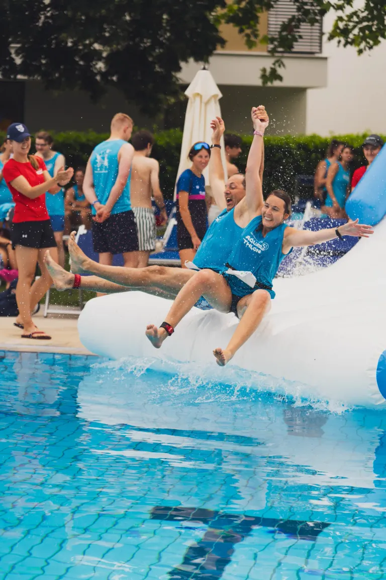 Eine Gruppe von Menschen in blauen Shirts und Shorts fährt auf einer Wasserrutsche, eine Person hält die Hand der anderen. Sie sind bei einer Pool-Veranstaltung.