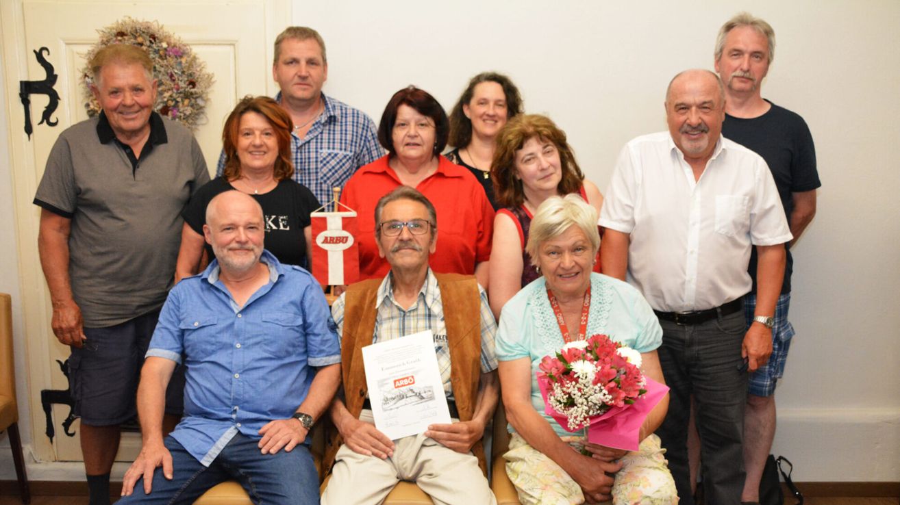 Eine Gruppe älterer Menschen ist für ein Foto versammelt. Ein Mann in der Mitte hält ein Zertifikat, während eine Frau neben ihm einen Blumenstrauß hält. Sie alle lächeln.