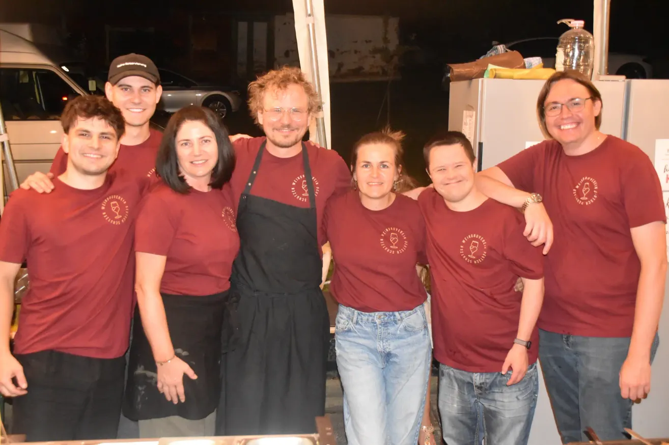 Eine Gruppe von Menschen in roten T-Shirts posiert für ein Foto. Eine Person trägt eine schwarze Schürze. Sie stehen draußen, wobei ein Kühlschrank und ein Auto im Hintergrund zu sehen sind.