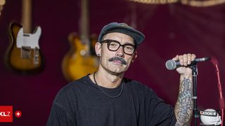 Ein Mann mit Schnauzbart und Brille, der eine Mütze trägt, singt in ein Mikrofon. Er steht vor einem lila Hintergrund mit Gitarren im Hintergrund.