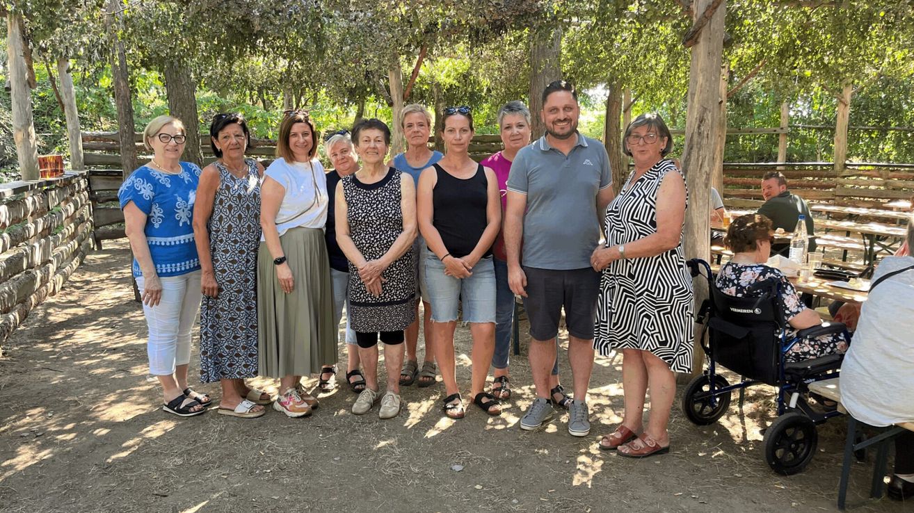 Eine Gruppe von Menschen steht unter einem Baumdach. Sie lächeln alle und posieren für ein Foto. Sie sind in Freizeitkleidung gekleidet und tragen Sandalen. Die Frau auf der rechten Seite ist im Rollstuhl. Die Schatten der Bäume fallen auf den Boden.