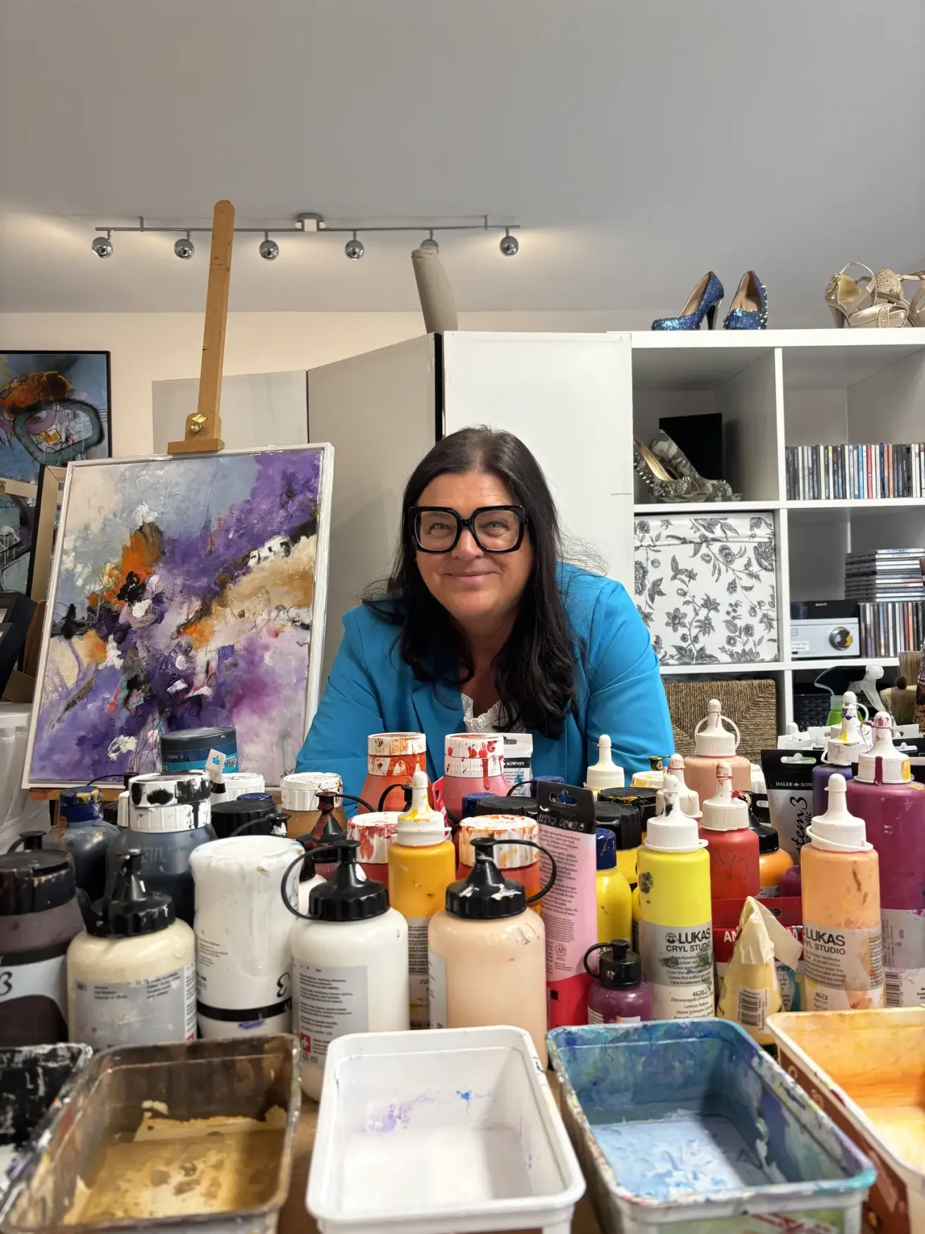 Eine Frau sitzt in einem Studio mit einer Vielzahl von Farbflaschen und einem Gemälde auf einem Staffelei. Sie trägt eine Brille und eine blaue Jacke. Hinter ihr stehen Regale mit hohen Schuhen, CDs und anderen Gegenständen.