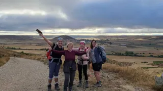 Vier Wanderer posieren für ein Foto auf einem Schotterweg, mit weitläufigen ländlichen Landschaften und Hügeln im Hintergrund. Die Frau in der Mitte hält eine Kamera.