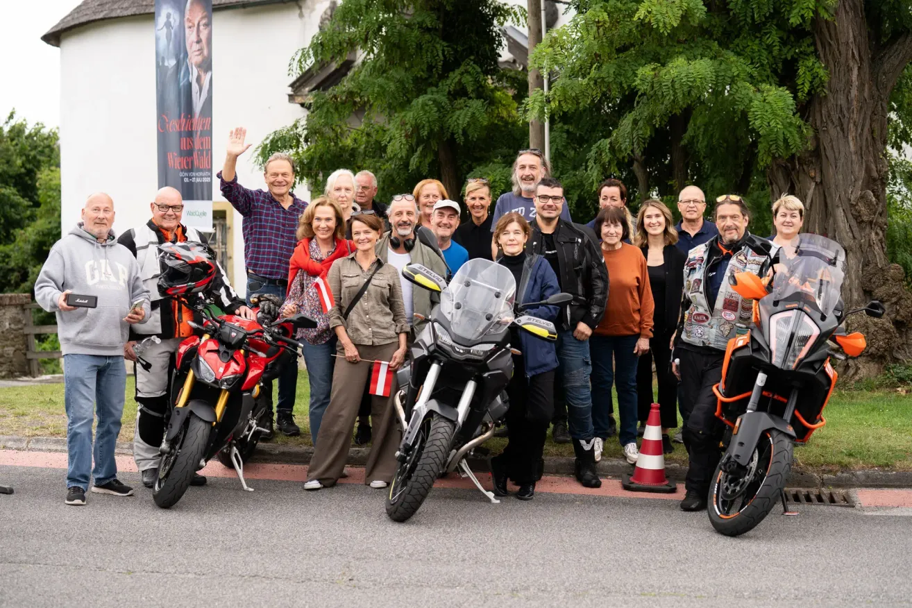 Eine Gruppe von Menschen steht auf der Straße, einige halten Motorräder, vor einem Gebäude mit einem Plakat. Sie tragen Jacken und manche tragen Helme. Sie posieren für ein Foto. Die Motorräder sind am Straßenrand geparkt. Es gibt Bäume und einen Verkehrskegel in der Umgebung.