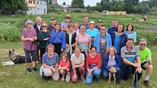 Eine Gruppe von Menschen, einschließlich Erwachsener und Kinder, posiert für ein Foto in einem Garten. Einige sitzen, andere stehen. Im Hintergrund befinden sich Gebäude, Bäume und ein klarer Himmel.