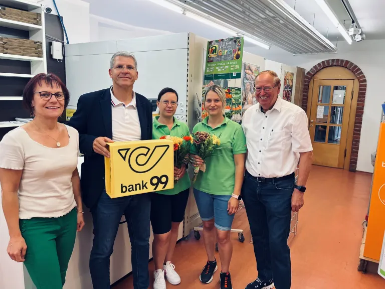 Fünf Menschen in grünen Shirts und mit Brillen stehen in einem Raum mit einer gelben Box und lächeln. Zwei Frauen halten Blumen. Ein Mann trägt einen schwarzen Anzug.