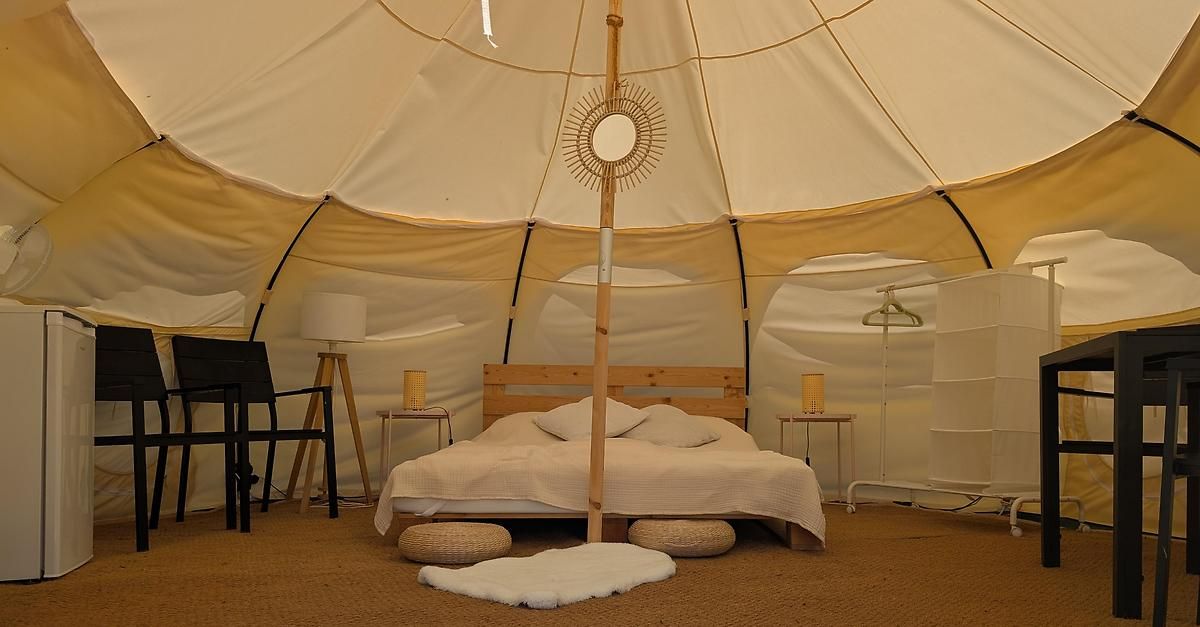Ein weißes Zelt mit einem Holzbett, zwei Kissen, einem weißen Pelzteppich und zwei kleinen Tischen mit Lampen. Es gibt auch einen Stuhl und eine Lampe auf einem Ständer.