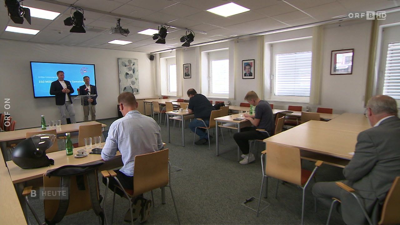 Ein Klassenzimmer mit Tischen und Stühlen, ein Mann steht an einem Rednerpult. Mehrere Männer sitzen an Tischen, einige mit Brillen, ein Mann mit Laptop. Bilderrahmen schmücken die Wände, und ein Monitor ist sichtbar.